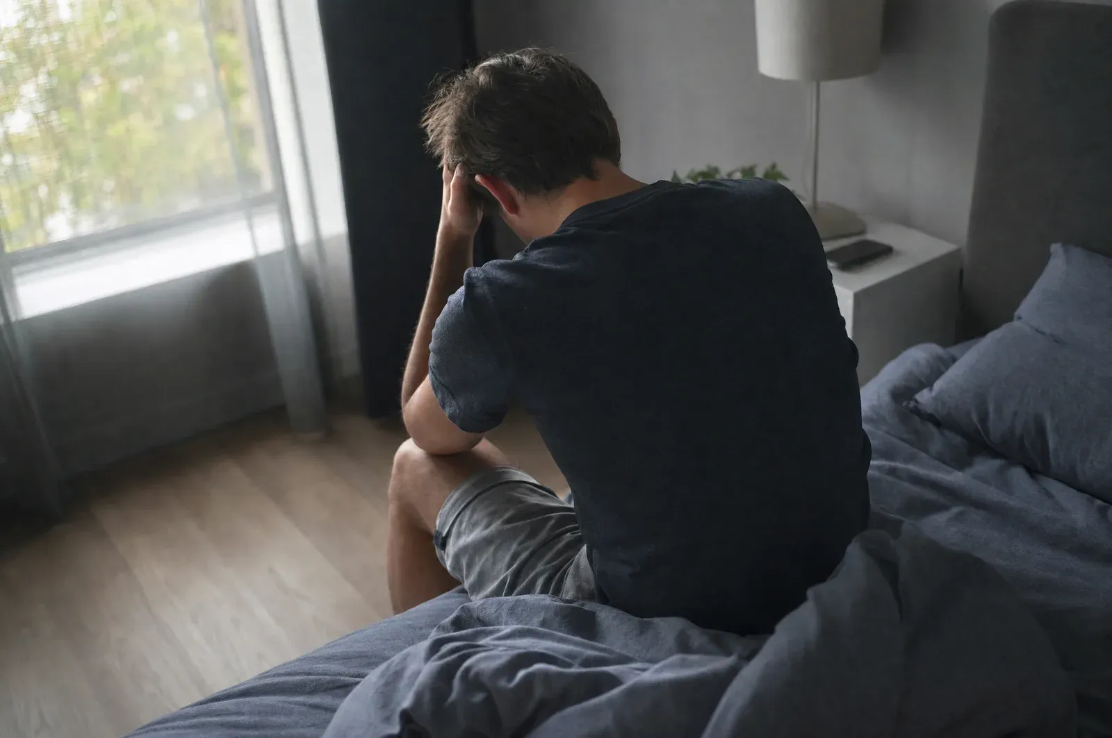 A person sitting on the edge of a bed in a dim room, one hand pressed to the side of their head.